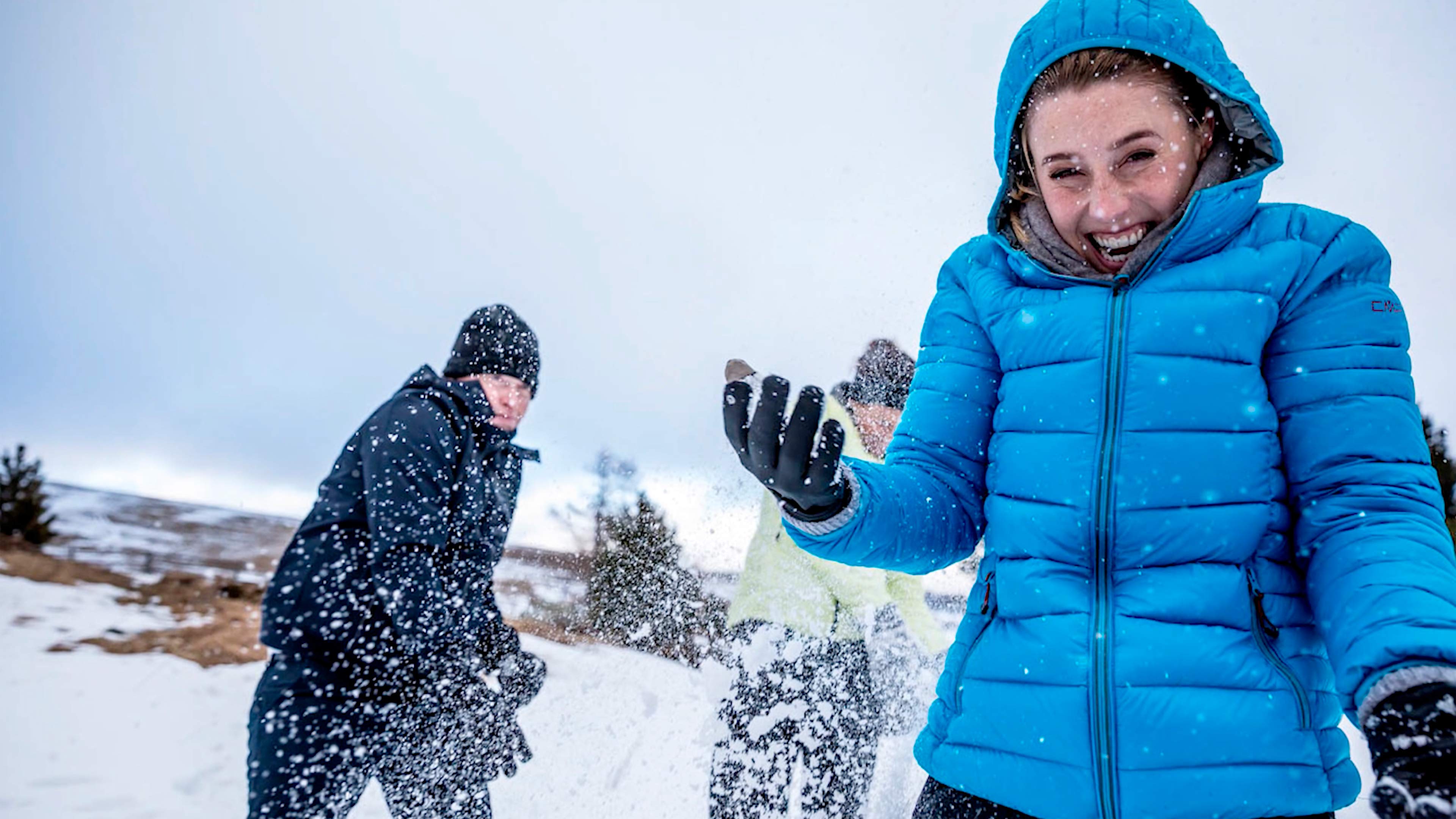 Drei Personen im Schnee, sie tragen Winterkleidung. Eine Frau im Vordergrund trägt eine blaue Daunenjacke, sie lacht in die Kamera. 